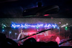 A nighttime scene through a vehicle's windshield shows a large illuminated sign reading 'Welcome To Cairo' in both English and Arabic. There are multiple lanes and vehicles passing through, with a toll booth visible. Bright lights illuminate the area, highlighting the structure's modern design.
