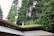 A modern building with a green roof is set against a backdrop of tall, lush evergreen trees. The architecture features angular lines and large glass windows, integrating the structure harmoniously with the surrounding natural landscape.