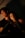 A young couple sits closely together in a dimly lit setting, with warm light casting soft shadows across their faces. The woman rests her head on the man's shoulder, conveying a sense of intimacy and comfort.