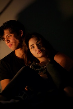 A young couple sits closely together in a dimly lit setting, with warm light casting soft shadows across their faces. The woman rests her head on the man's shoulder, conveying a sense of intimacy and comfort.