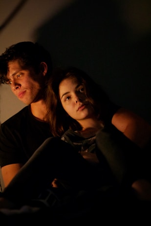 A young couple sits closely together in a dimly lit setting, with warm light casting soft shadows across their faces. The woman rests her head on the man's shoulder, conveying a sense of intimacy and comfort.