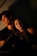 A young couple sits closely together in a dimly lit setting, with warm light casting soft shadows across their faces. The woman rests her head on the man's shoulder, conveying a sense of intimacy and comfort.
