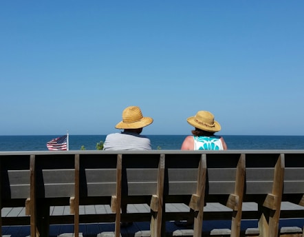 Two people wearing straw hats sit on a bench looking out at the ocean, with a clear blue sky overhead. An American flag is visible to the left, gently waving in the breeze.