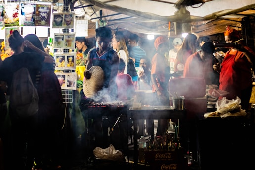 A bustling street food stall is filled with people. A person is tending to a grill, fanning the flames with a woven fan to cook skewers of meat. Smoke rises from the grill, mingling with the lights of the brightly decorated stand. Various food and drink images are displayed on the stall, with customers in casual outfits enjoying the lively atmosphere.