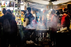 A bustling street food stall is filled with people. A person is tending to a grill, fanning the flames with a woven fan to cook skewers of meat. Smoke rises from the grill, mingling with the lights of the brightly decorated stand. Various food and drink images are displayed on the stall, with customers in casual outfits enjoying the lively atmosphere.