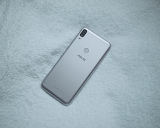 A silver-colored smartphone with dual rear cameras and a fingerprint sensor is placed on a soft, textured fabric surface. The brand logo is visible on the phone's back.