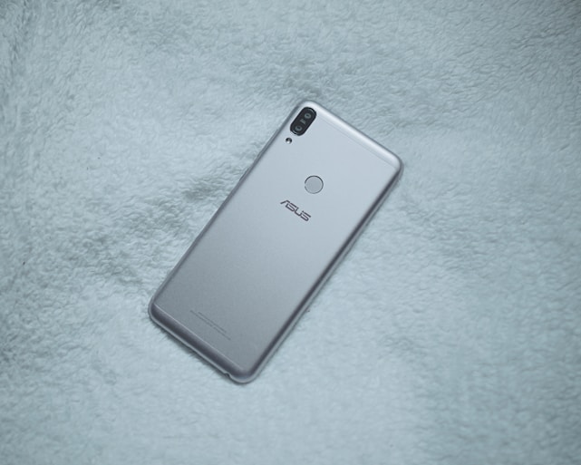 A silver-colored smartphone with dual rear cameras and a fingerprint sensor is placed on a soft, textured fabric surface. The brand logo is visible on the phone's back.