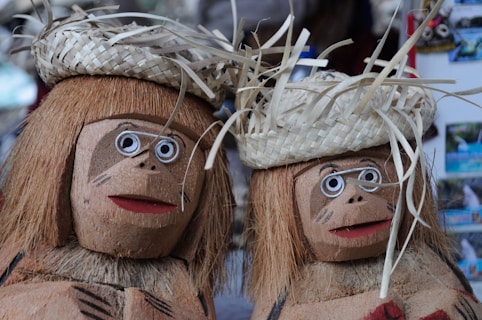 Two handcrafted figures made from coconut shells and fibers, featuring facial designs and straw hats. Each figure has large white circular eyes with black centers, a smiling mouth, and decorative markings on the face. The hats appear to be woven from natural materials.