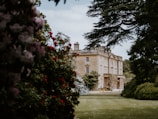 A large, stately home with classic architecture is surrounded by lush gardens. The building is framed by trees and colorful flowering shrubs, creating a picturesque and serene setting.