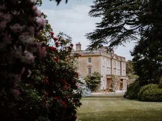 A large, stately home with classic architecture is surrounded by lush gardens. The building is framed by trees and colorful flowering shrubs, creating a picturesque and serene setting.