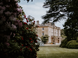 A large, stately home with classic architecture is surrounded by lush gardens. The building is framed by trees and colorful flowering shrubs, creating a picturesque and serene setting.