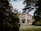 A large, stately home with classic architecture is surrounded by lush gardens. The building is framed by trees and colorful flowering shrubs, creating a picturesque and serene setting.