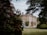 A large, stately home with classic architecture is surrounded by lush gardens. The building is framed by trees and colorful flowering shrubs, creating a picturesque and serene setting.