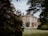 A large, stately home with classic architecture is surrounded by lush gardens. The building is framed by trees and colorful flowering shrubs, creating a picturesque and serene setting.