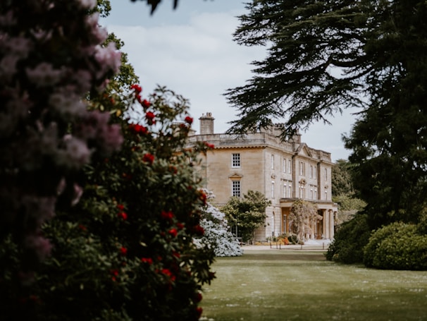 A large, stately home with classic architecture is surrounded by lush gardens. The building is framed by trees and colorful flowering shrubs, creating a picturesque and serene setting.