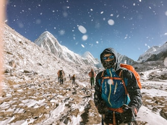 Several mountaineers are trekking through a snowy mountainous landscape. The scene features high, snow-capped peaks under a clear blue sky. The climbers are dressed in winter gear, with backpacks and protective outerwear. Snowflakes can be seen in the air, adding to the cold and adventurous atmosphere.