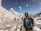 Several mountaineers are trekking through a snowy mountainous landscape. The scene features high, snow-capped peaks under a clear blue sky. The climbers are dressed in winter gear, with backpacks and protective outerwear. Snowflakes can be seen in the air, adding to the cold and adventurous atmosphere.