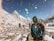 Several mountaineers are trekking through a snowy mountainous landscape. The scene features high, snow-capped peaks under a clear blue sky. The climbers are dressed in winter gear, with backpacks and protective outerwear. Snowflakes can be seen in the air, adding to the cold and adventurous atmosphere.