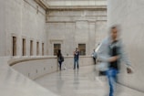 An architectural interior space with high ceilings and large stone walls. A person is centered in the background, appearing to take a photo or video. Another person walks past in the foreground, blurred from motion, while two other individuals are visible in the background. The setting appears calm with an elegant, minimalist design.