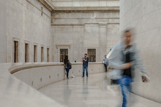 An architectural interior space with high ceilings and large stone walls. A person is centered in the background, appearing to take a photo or video. Another person walks past in the foreground, blurred from motion, while two other individuals are visible in the background. The setting appears calm with an elegant, minimalist design.