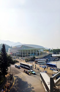 A large, modern stadium is situated near a busy road with several buses and a taxi in the foreground. People are walking along the sidewalks and the area is surrounded by trees and hills. The architecture of the stadium is contemporary with a white facade and glass panels. The sky is clear with natural daylight illuminating the scene.