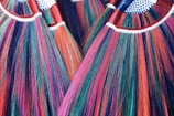 A close-up view of several vibrant, multicolored brooms with bristles in shades of red, purple, green, and blue. The handles are wrapped in red tape and have a pattern of white and blue checkered fabric at the top.