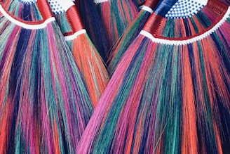 A close-up view of several vibrant, multicolored brooms with bristles in shades of red, purple, green, and blue. The handles are wrapped in red tape and have a pattern of white and blue checkered fabric at the top.