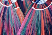 A close-up view of several vibrant, multicolored brooms with bristles in shades of red, purple, green, and blue. The handles are wrapped in red tape and have a pattern of white and blue checkered fabric at the top.