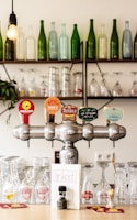 A bar setting with a row of colorful beer taps prominently displayed in the foreground. Glasses of various shapes and sizes are neatly arranged below the taps. Shelves in the background hold a variety of empty bottles in shades of green and clear glass, adding to the decor. A single light bulb illuminates the scene with a warm glow, contributing to a cozy atmosphere.