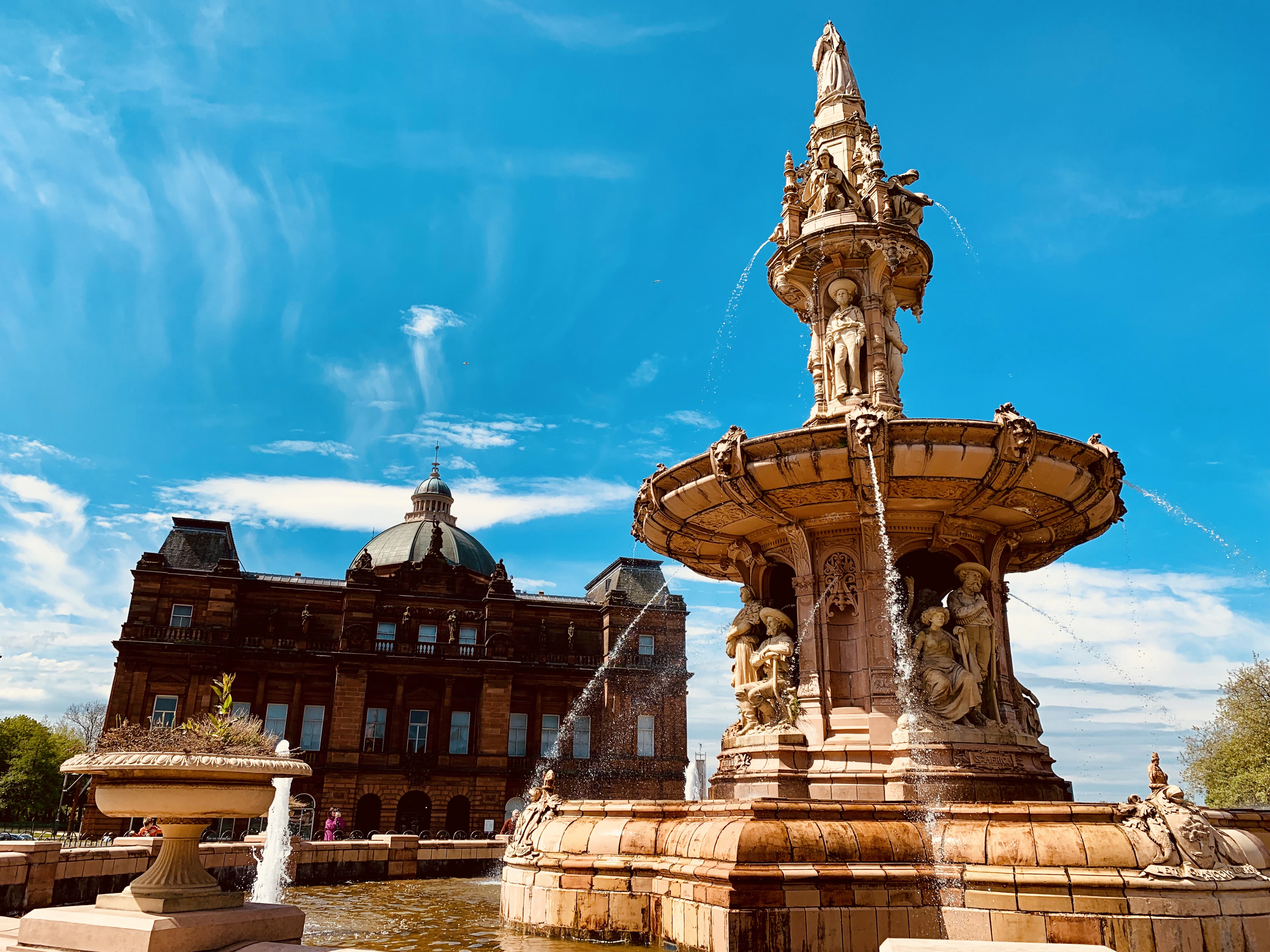 The terracotta Doulton Fountain on Glasgow Green in full flow, its tiered figures and basins catching the sun, with the red sandstone People's Palace and its glass Winter Gardens dome behind under a vivid blue sky.