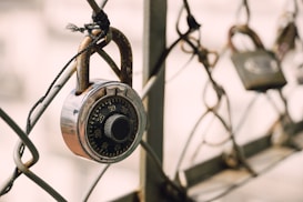 A close-up of a metallic combination padlock attached to a wire fence. The lock's numbers are clearly visible, and the focus is sharp on the lock, with a background featuring other blurred locks.