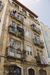 A multi-story residential building with ornate balconies and patterned facade. Clothes are hanging to dry on several balconies while potted plants add a touch of greenery. The building shows signs of aging but maintains a charming character.