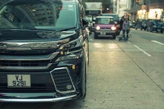 A street scene featuring a shiny black vehicle prominently in the foreground with a visible license plate. In the background, another smaller car is parked, and a person is walking with a stroller. The setting is urban with buildings lining the street and other vehicles visible in the distance.
