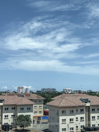 A residential area with several beige-colored apartment buildings with red-tiled roofs. In the background, there are taller buildings and lush greenery. The sky overhead is mostly clear with some wispy clouds.
