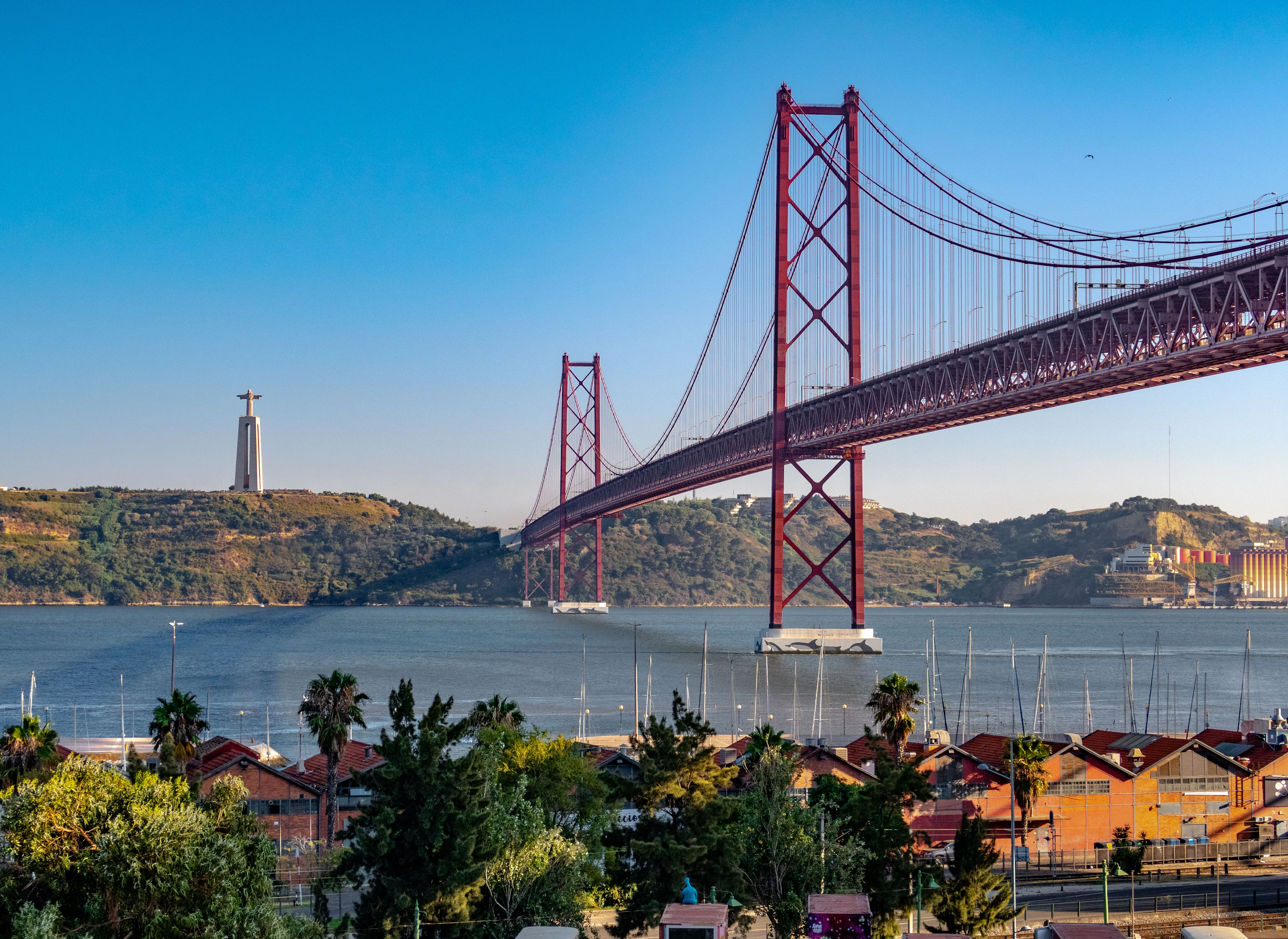 The red steel Ponte 25 de Abril spanning the Tagus in bright afternoon sun, with the Cristo Rei statue on the green hillside beyond and terracotta rooftops along the Lisbon shore