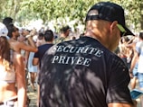 A man wearing a black t-shirt with 'Securite Privee' printed on the back is standing in a crowded outdoor setting. Many people are visible in the background, some holding drinks. The environment suggests a casual, possibly festive atmosphere with sunlit trees casting dappled shadows.