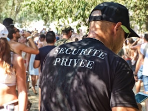 A man wearing a black t-shirt with 'Securite Privee' printed on the back is standing in a crowded outdoor setting. Many people are visible in the background, some holding drinks. The environment suggests a casual, possibly festive atmosphere with sunlit trees casting dappled shadows.