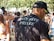 A man wearing a black t-shirt with 'Securite Privee' printed on the back is standing in a crowded outdoor setting. Many people are visible in the background, some holding drinks. The environment suggests a casual, possibly festive atmosphere with sunlit trees casting dappled shadows.