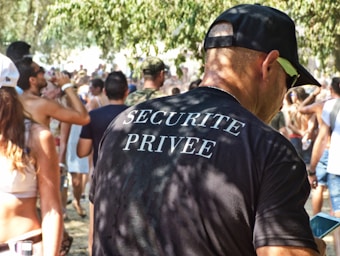 A man wearing a black t-shirt with 'Securite Privee' printed on the back is standing in a crowded outdoor setting. Many people are visible in the background, some holding drinks. The environment suggests a casual, possibly festive atmosphere with sunlit trees casting dappled shadows.