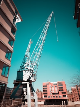 A large crane stands prominently between two buildings, with its boom extending upward against a clear turquoise sky. The crane has a metallic structure and is situated on a platform, surrounded by modern apartment buildings in an urban setting.