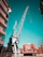 A large crane stands prominently between two buildings, with its boom extending upward against a clear turquoise sky. The crane has a metallic structure and is situated on a platform, surrounded by modern apartment buildings in an urban setting.