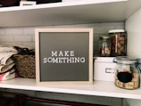 A neatly organized shelf displays a letter board with the phrase 'MAKE SOMETHING' in white letters on a gray background. Surrounding the board are various crafting supplies including woven baskets, quilts, glass jars filled with wooden stamps, and a white storage box. The background features a light gray brick wall.