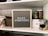 A neatly organized shelf displays a letter board with the phrase 'MAKE SOMETHING' in white letters on a gray background. Surrounding the board are various crafting supplies including woven baskets, quilts, glass jars filled with wooden stamps, and a white storage box. The background features a light gray brick wall.