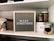 A neatly organized shelf displays a letter board with the phrase 'MAKE SOMETHING' in white letters on a gray background. Surrounding the board are various crafting supplies including woven baskets, quilts, glass jars filled with wooden stamps, and a white storage box. The background features a light gray brick wall.