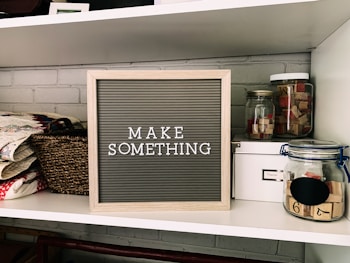 A neatly organized shelf displays a letter board with the phrase 'MAKE SOMETHING' in white letters on a gray background. Surrounding the board are various crafting supplies including woven baskets, quilts, glass jars filled with wooden stamps, and a white storage box. The background features a light gray brick wall.