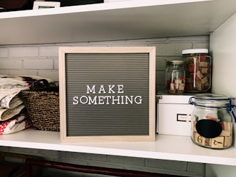 A neatly organized shelf displays a letter board with the phrase 'MAKE SOMETHING' in white letters on a gray background. Surrounding the board are various crafting supplies including woven baskets, quilts, glass jars filled with wooden stamps, and a white storage box. The background features a light gray brick wall.