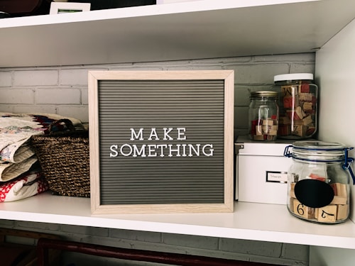 A neatly organized shelf displays a letter board with the phrase 'MAKE SOMETHING' in white letters on a gray background. Surrounding the board are various crafting supplies including woven baskets, quilts, glass jars filled with wooden stamps, and a white storage box. The background features a light gray brick wall.