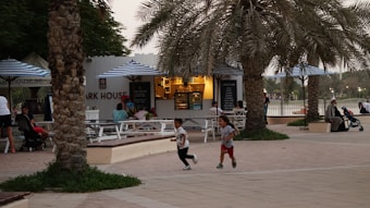 A park scene with children playing in the foreground near palm trees and a small caf&eacute; or food stand with blue-and-white striped umbrellas. People are seated at picnic tables, and a man with a stroller is also visible. The atmosphere is relaxed and casual.