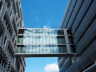 A glass-walled sky bridge connects two modern office buildings, reflecting the blue sky and clouds. The buildings are composed of vertical and horizontal lines with a sleek and contemporary design.