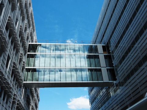 A glass-walled sky bridge connects two modern office buildings, reflecting the blue sky and clouds. The buildings are composed of vertical and horizontal lines with a sleek and contemporary design.